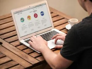 Person typing on laptop outdoors, illustrating remote work lifestyle with coffee at a wooden table.